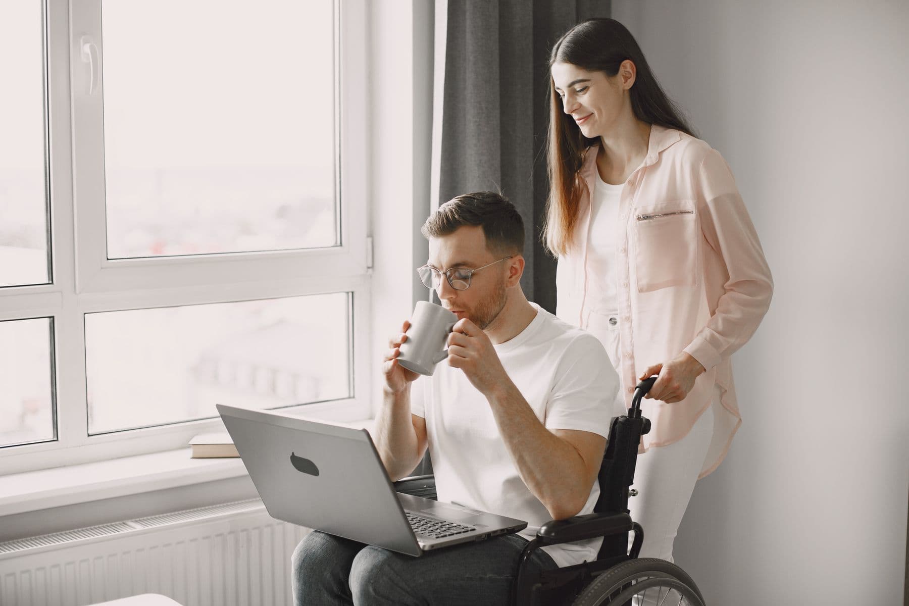 A man using a laptop in his wheelchair at home, with someone beside him looking at the screen